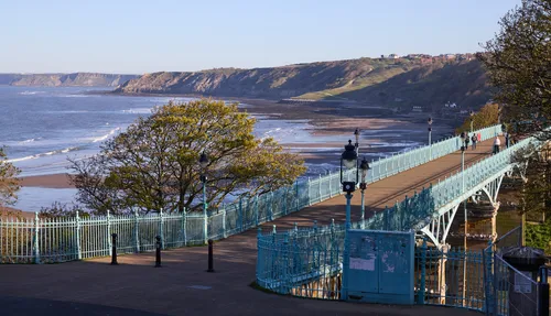 Scarborough cliffs and bridge This landscape photograph shows the Scarborough cliffs and the ornate iron bridge stretching across the promenade in Yorkshire. The image was taken in the late afternoon during spring, with sunlight casting gentle shadows and illuminating the fresh green foliage of the trees. The cliffs of Scarborough dominate the background, where waves gently lap against the sandy shore and the open sea extends outwards. People can be seen walking along the bridge, which features decorative street lamps and intricate railings, reflecting classic Victorian architectural style. The location is a popular coastal destination in Yorkshire, and the photograph captures the scenic beauty and tranquil atmosphere of Scarborough’s shoreline and cliffs during the springtime.